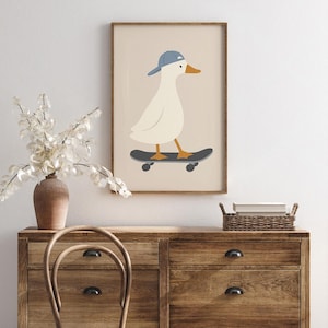 May include: A framed art print featuring a cartoon duck wearing a blue baseball cap and riding a skateboard. The duck is white with an orange beak and feet. The skateboard is black with gray wheels. The background is a light beige color.
