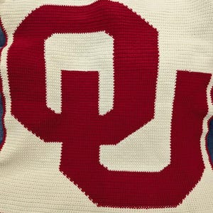 May include: A handmade crochet blanket featuring the letter "OU" in red against a white background. The blanket is made with a tight stitch, showcasing the team's colors. The blanket is rectangular and appears to be on a denim surface.