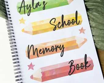 Könnte beinhalten: Ein weißes Spiralnotizbuch mit einem bunten Bleistiftdesign. Das Notizbuch trägt den Text "Ayla's School Memory Book" auf dem Cover.