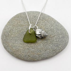 May include: A silver necklace with a green sea glass pendant and a small seashell charm. The necklace is presented on a grey stone. The sea glass is triangular. The background is white.