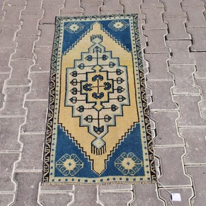 Puede incluir: Una alfombra pequeña y rectangular con un patrón geométrico en tonos azules, amarillos y marrones. La alfombra tiene un borde con flecos.
