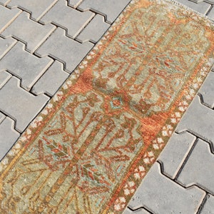 May include: A vintage runner rug with a muted green, orange, and white geometric pattern. The rug is laid out on a gray concrete patio.