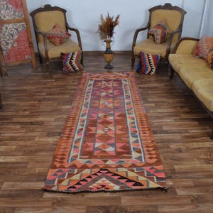 May include: A long rectangular runner rug featuring a geometric pattern in shades of orange, pink, brown, and cream. The rug displays repeating triangular and diamond motifs arranged in a central panel with a decorative border along the edges.