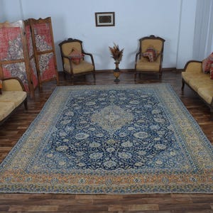 K&ouml;nnte beinhalten: Ein gro&szlig;er, rechteckiger, blau-beiger Perserteppich mit einem komplizierten Blumenmuster dominiert den Raum. Der Teppich ist von antiken M&ouml;beln umgeben, darunter zwei Sofas, zwei Sessel und ein Paravent. Der Raum hat eine warme, einladende Atmosph&auml;re.