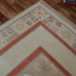 May include: Cream-colored area rug with a geometric design. The rug features a central cream field bordered by a rust-colored frame and decorative patterns in brown and rust. The rug is placed on a wooden floor.