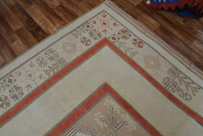 May include: Cream-colored area rug with a geometric design. The rug features a central cream field bordered by a rust-colored frame and decorative patterns in brown and rust. The rug is placed on a wooden floor.