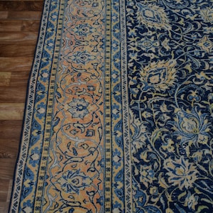 K&ouml;nnte beinhalten: Ein Abschnitt eines verzierten Teppichs mit detailliertem Blumenmuster. Der Teppich hat ein marineblaues Feld mit cremefarbenen und hellblauen Blumenmotiven. Ein Rand mit einem sich wiederholenden Blumenmuster in Orange-, Blau- und Cremet&ouml;nen rahmt den Teppich ein.