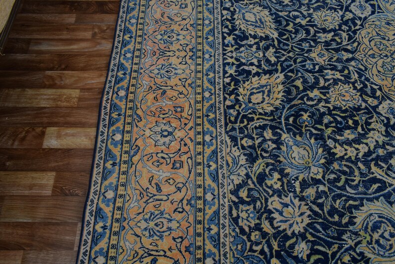 K&ouml;nnte beinhalten: Ein Abschnitt eines verzierten Teppichs mit detailliertem Blumenmuster. Der Teppich hat ein marineblaues Feld mit cremefarbenen und hellblauen Blumenmotiven. Ein Rand mit einem sich wiederholenden Blumenmuster in Orange-, Blau- und Cremet&ouml;nen rahmt den Teppich ein.