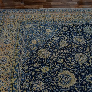 K&ouml;nnte beinhalten: Eckansicht eines gro&szlig;en, verzierten Teppichs mit marineblauem Feld und cremefarbenem Rand. Der Teppich weist ein Blumenmuster mit komplizierten Details in Blau-, Creme- und Goldt&ouml;nen auf. Der Teppich liegt auf einem Holzboden.
