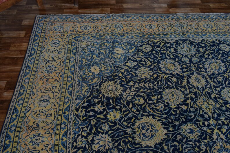 K&ouml;nnte beinhalten: Eckansicht eines gro&szlig;en, verzierten Teppichs mit marineblauem Feld und cremefarbenem Rand. Der Teppich weist ein Blumenmuster mit komplizierten Details in Blau-, Creme- und Goldt&ouml;nen auf. Der Teppich liegt auf einem Holzboden.