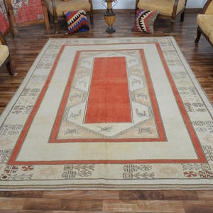 May include: A large rectangular rug with a cream-colored field and a central orange panel. The rug features a border with geometric patterns in orange, brown, and beige. The rug is placed on a wooden floor.