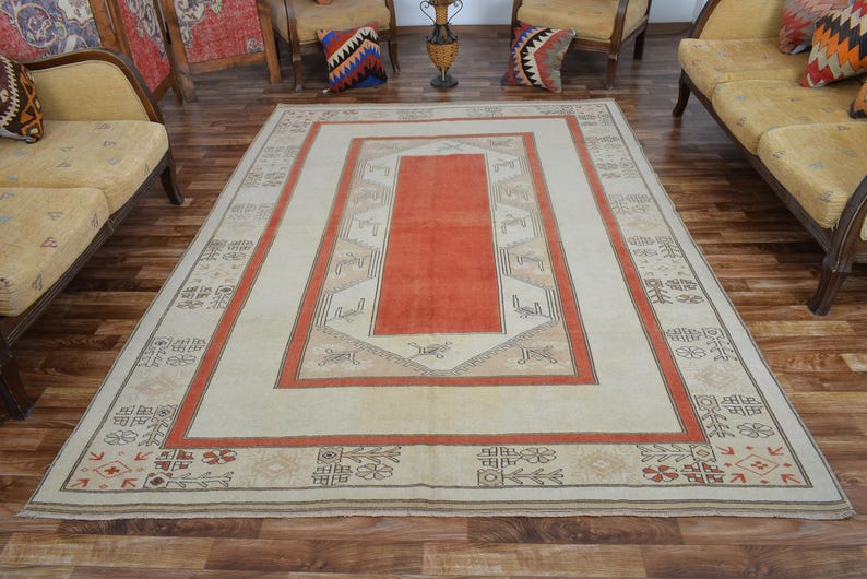 May include: A large rectangular rug with a cream-colored field and a central orange panel. The rug features a border with geometric patterns in orange, brown, and beige. The rug is placed on a wooden floor.
