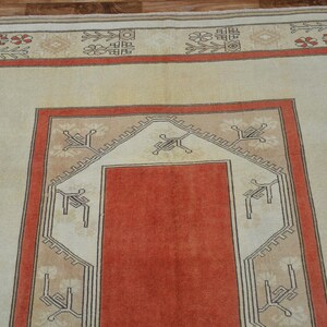 May include: A rectangular rug with a central orange field, framed by a beige border with geometric patterns in red and black. The rug's design features a symmetrical layout with repeating motifs.