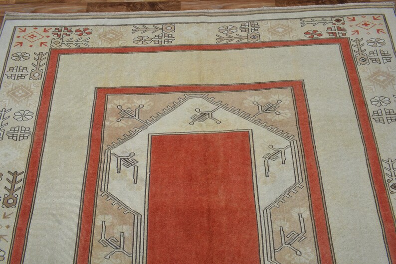 May include: A rectangular rug with a central orange field, framed by a beige border with geometric patterns in red and black. The rug's design features a symmetrical layout with repeating motifs.