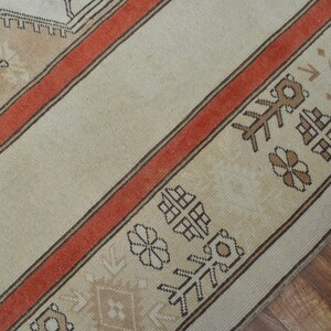 May include: Close-up of a beige and rust-colored rug with geometric patterns. The rug features a central beige field with a rust-colored border and decorative motifs. The border includes repeating floral and geometric designs.