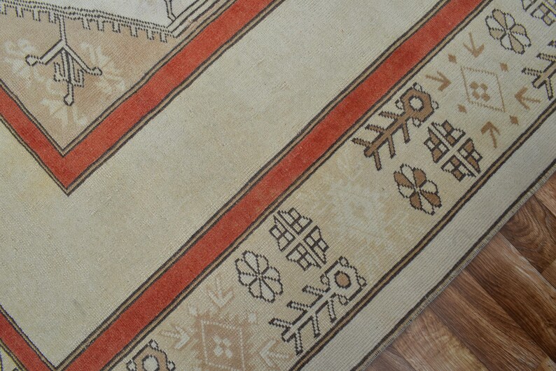 May include: Close-up of a beige and rust-colored rug with geometric patterns. The rug features a central beige field with a rust-colored border and decorative motifs. The border includes repeating floral and geometric designs.