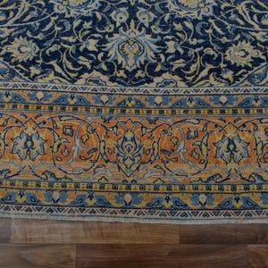 K&ouml;nnte beinhalten: Nahaufnahme eines Teils eines Orientteppichs mit detailliertem Blumenmuster. Der Teppich hat ein marineblaues Feld mit cremefarbenen und hellblauen Blumenmotiven. Ein Rand aus orangefarbenen, blauen und cremefarbenen Blumendesigns umrahmt das zentrale Muster. Der Teppich liegt auf einem Holzboden.