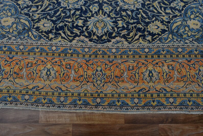 K&ouml;nnte beinhalten: Nahaufnahme eines Teils eines Orientteppichs mit detailliertem Blumenmuster. Der Teppich hat ein marineblaues Feld mit cremefarbenen und hellblauen Blumenmotiven. Ein Rand aus orangefarbenen, blauen und cremefarbenen Blumendesigns umrahmt das zentrale Muster. Der Teppich liegt auf einem Holzboden.