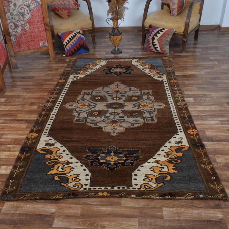 May include: A rectangular, brown area rug with an intricate central medallion design in shades of gray, beige, and yellow. The rug features a border with floral and geometric patterns, creating a traditional aesthetic. The rug is placed on a wooden floor.