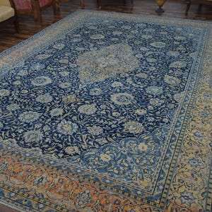 K&ouml;nnte beinhalten: Gro&szlig;er rechteckiger Teppich mit marineblauem Feld und aufwendigen Blumenmustern in Hellblau und Creme. Der Rand ist eine Mischung aus Orange, Gelb und Blau, die einen auff&auml;lligen Kontrast erzeugt. Der Teppich liegt auf einem Holzboden.