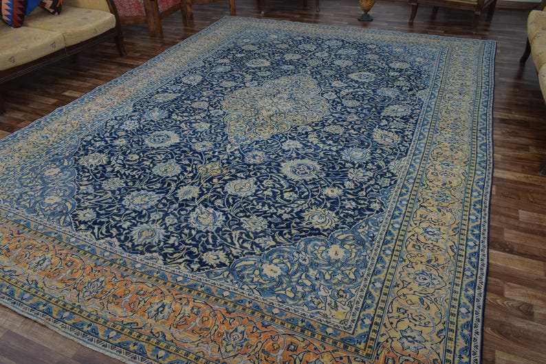 K&ouml;nnte beinhalten: Gro&szlig;er rechteckiger Teppich mit marineblauem Feld und aufwendigen Blumenmustern in Hellblau und Creme. Der Rand ist eine Mischung aus Orange, Gelb und Blau, die einen auff&auml;lligen Kontrast erzeugt. Der Teppich liegt auf einem Holzboden.