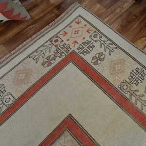 May include: Close-up of a beige and rust-colored area rug with geometric patterns. The rug features a central diamond design framed by a rust-colored border and decorative motifs. The rug is placed on a wooden floor.