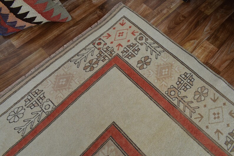 May include: Close-up of a beige and rust-colored area rug with geometric patterns. The rug features a central diamond design framed by a rust-colored border and decorative motifs. The rug is placed on a wooden floor.