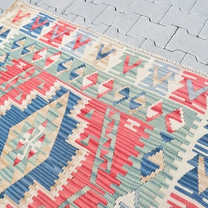 May include: A traditional Turkish kilim rug with a geometric pattern in shades of red, blue, green, and beige. The rug has a fringed edge and is woven with a variety of textures.