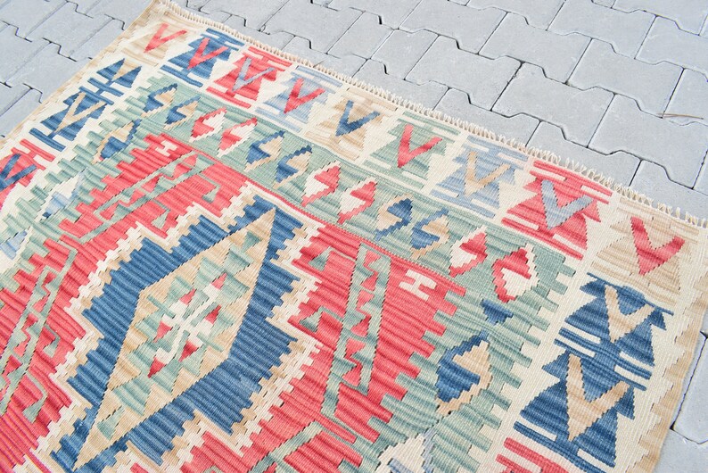 May include: A traditional Turkish kilim rug with a geometric pattern in shades of red, blue, green, and beige. The rug has a fringed edge and is woven with a variety of textures.