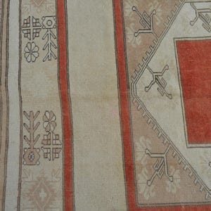 May include: Close-up of a section of a vintage rug with a geometric design. The rug features a central red field framed by a beige border with intricate patterns. Vertical stripes in beige and red add to the design. The rug is likely made of wool.