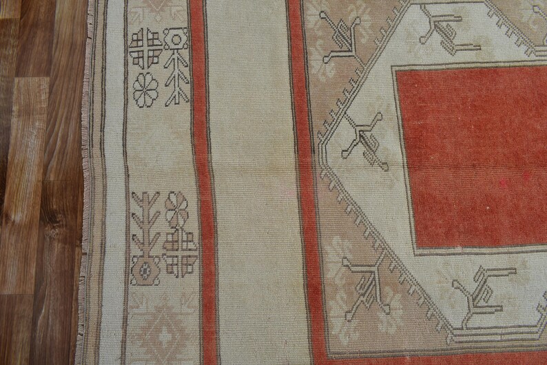 May include: Close-up of a section of a vintage rug with a geometric design. The rug features a central red field framed by a beige border with intricate patterns. Vertical stripes in beige and red add to the design. The rug is likely made of wool.