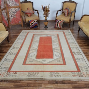 May include: A large rectangular rug with a central orange panel and a cream-colored border. The border features geometric patterns in shades of orange, beige, and brown. The rug is placed on a wooden floor, with antique chairs and decorative items in the background.
