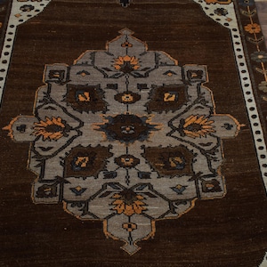 May include: A rectangular brown rug with a central medallion design in grey, brown, and orange. The rug features a border with floral and geometric patterns. The rug is placed on a wooden floor.