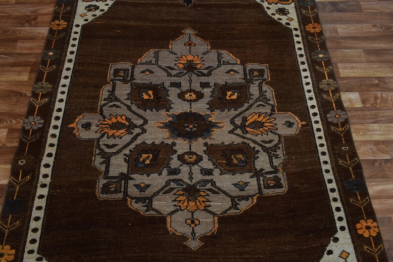 May include: A rectangular brown rug with a central medallion design in grey, brown, and orange. The rug features a border with floral and geometric patterns. The rug is placed on a wooden floor.