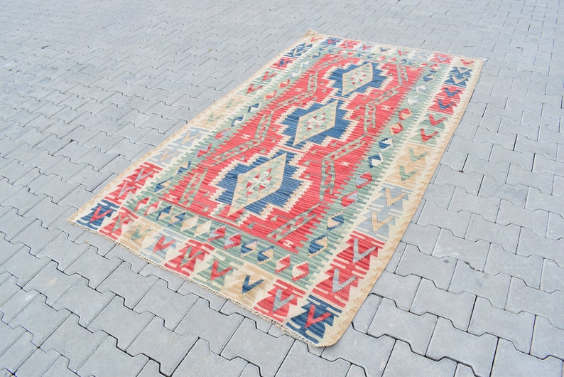 May include: A traditional Turkish kilim rug with a geometric pattern in shades of red, blue, green, and beige. The rug is woven with a flat weave and has a fringed edge.