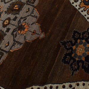 May include: Close-up of a brown and grey patterned rug. The rug features geometric designs in shades of brown, grey, and orange. The design includes floral and geometric shapes, with a border of small white dots.