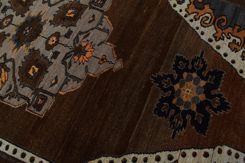 May include: Close-up of a brown and grey patterned rug. The rug features geometric designs in shades of brown, grey, and orange. The design includes floral and geometric shapes, with a border of small white dots.