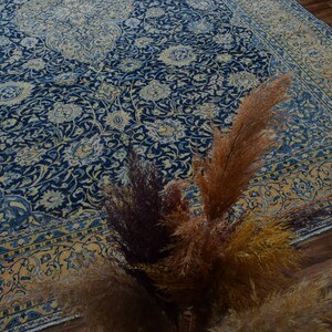 K&ouml;nnte beinhalten: Ein gro&szlig;er, verzierter Teppich mit marineblauem Feld und cremefarbenem Blumenmuster. Der Teppich hat einen cremefarbenen und goldenen Rand. Getrocknete Ziergr&auml;ser in Braun- und Goldt&ouml;nen befinden sich im Vordergrund.
