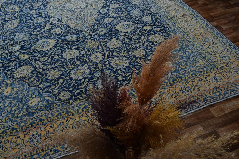 K&ouml;nnte beinhalten: Ein gro&szlig;er, verzierter Teppich mit marineblauem Feld und cremefarbenem Blumenmuster. Der Teppich hat einen cremefarbenen und goldenen Rand. Getrocknete Ziergr&auml;ser in Braun- und Goldt&ouml;nen befinden sich im Vordergrund.