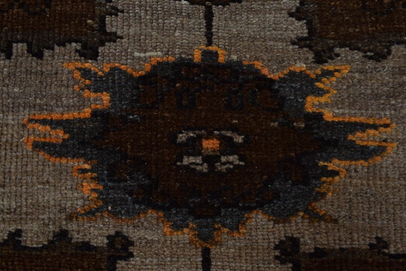 May include: Close-up of a section of a rug with a repeating pattern. The rug features a neutral beige background with dark brown and grey floral-like shapes. The edges of the shapes are outlined in orange.