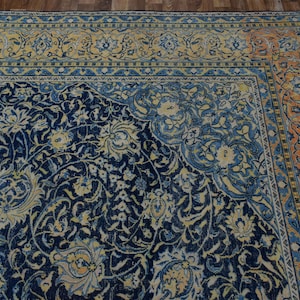 K&ouml;nnte beinhalten: Eckansicht eines detaillierten, verzierten Teppichs mit marineblauem Feld und floralen Mustern in Creme und Hellblau. Der Rand weist ein sich wiederholendes Design in Orange- und Blaut&ouml;nen auf. Der Teppich liegt auf einem Holzboden.