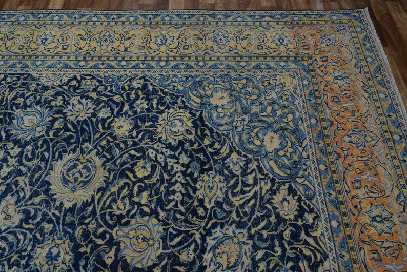 K&ouml;nnte beinhalten: Eckansicht eines detaillierten, verzierten Teppichs mit marineblauem Feld und floralen Mustern in Creme und Hellblau. Der Rand weist ein sich wiederholendes Design in Orange- und Blaut&ouml;nen auf. Der Teppich liegt auf einem Holzboden.