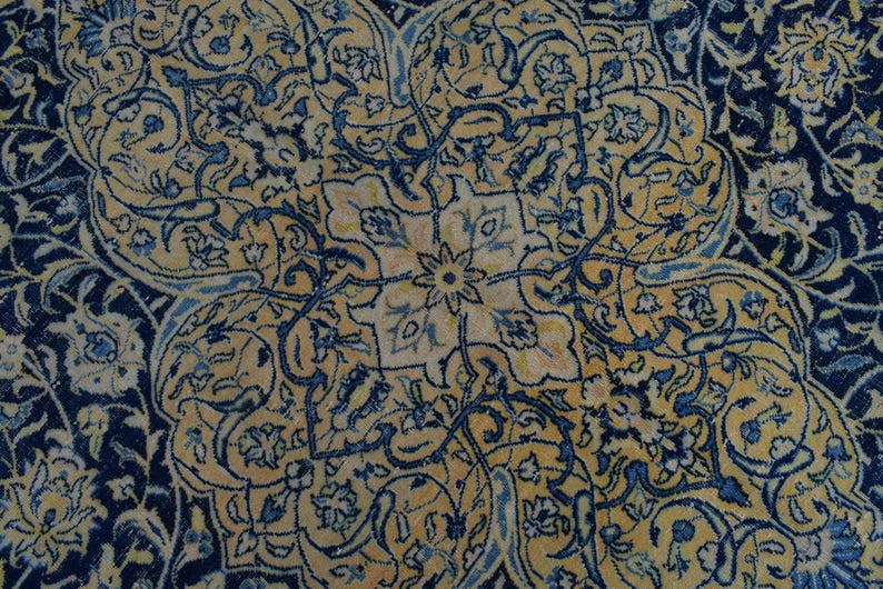 K&ouml;nnte beinhalten: Nahaufnahme eines verzierten Teppichs mit einem zentralen floralen Medaillon. Der Teppich hat einen marineblauen Rand und komplizierte Muster in Gold- und Blaut&ouml;nen. Das Design umfasst florale und arabeske Motive.