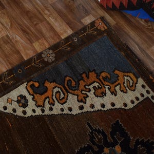 May include: A section of a brown rug with a blue and cream border. The rug features an intricate design with orange and brown patterns. The rug is placed on a wooden floor.