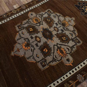 May include: A brown area rug with a central geometric design in grey, brown, and orange. The rug has a border with floral and geometric patterns in white, brown, and orange. The rug is on a wooden floor.