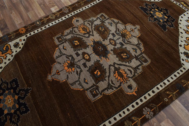 May include: A brown area rug with a central geometric design in grey, brown, and orange. The rug has a border with floral and geometric patterns in white, brown, and orange. The rug is on a wooden floor.