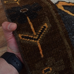 May include: Close-up of a section of a rug with a dark brown and grey background. The rug features a repeating pattern of orange and black geometric shapes. The rug is held by a person wearing a black watch.