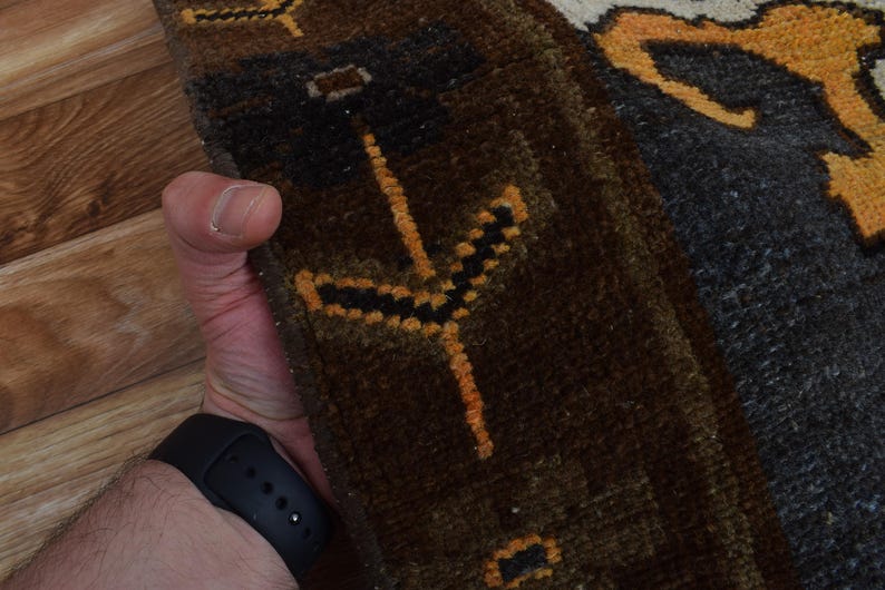 May include: Close-up of a section of a rug with a dark brown and grey background. The rug features a repeating pattern of orange and black geometric shapes. The rug is held by a person wearing a black watch.