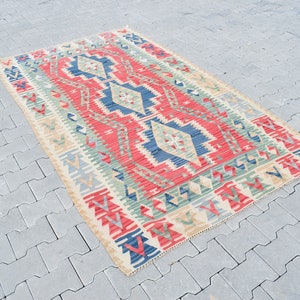 May include: A traditional Turkish kilim rug with a geometric pattern in shades of red, blue, green, and beige. The rug has a cream-colored background and is woven with intricate details.