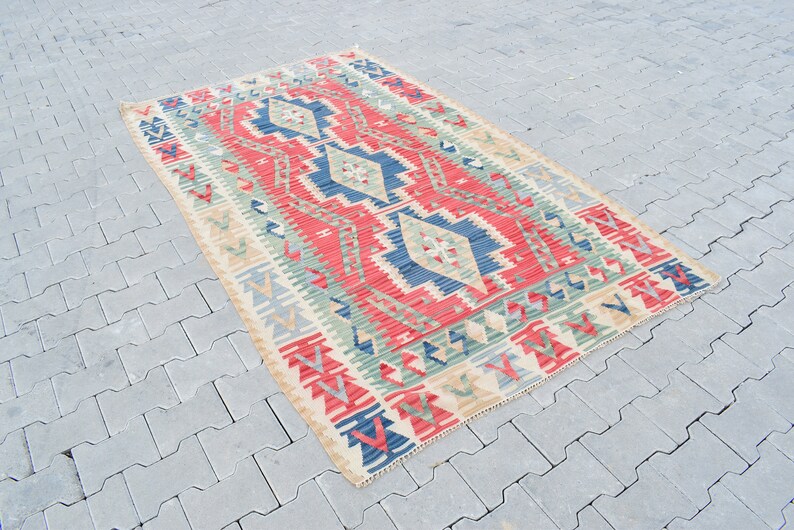 May include: A traditional Turkish kilim rug with a geometric pattern in shades of red, blue, green, and beige. The rug has a cream-colored background and is woven with intricate details.